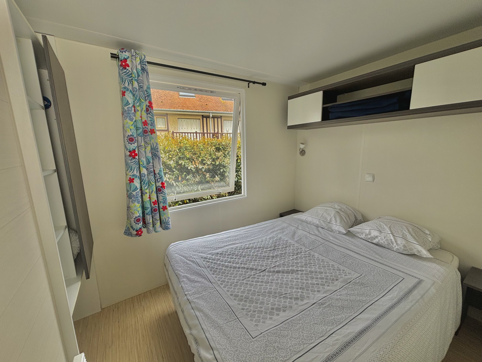Glamping-slaapkamer met tweepersoonsbed, kleurrijk gordijn, groot raam en uitzicht op groen en gebouw.