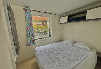 Dormitorio de glamping con cama doble, cortina floral, ventana grande y vista exterior de vegetación y edificio.