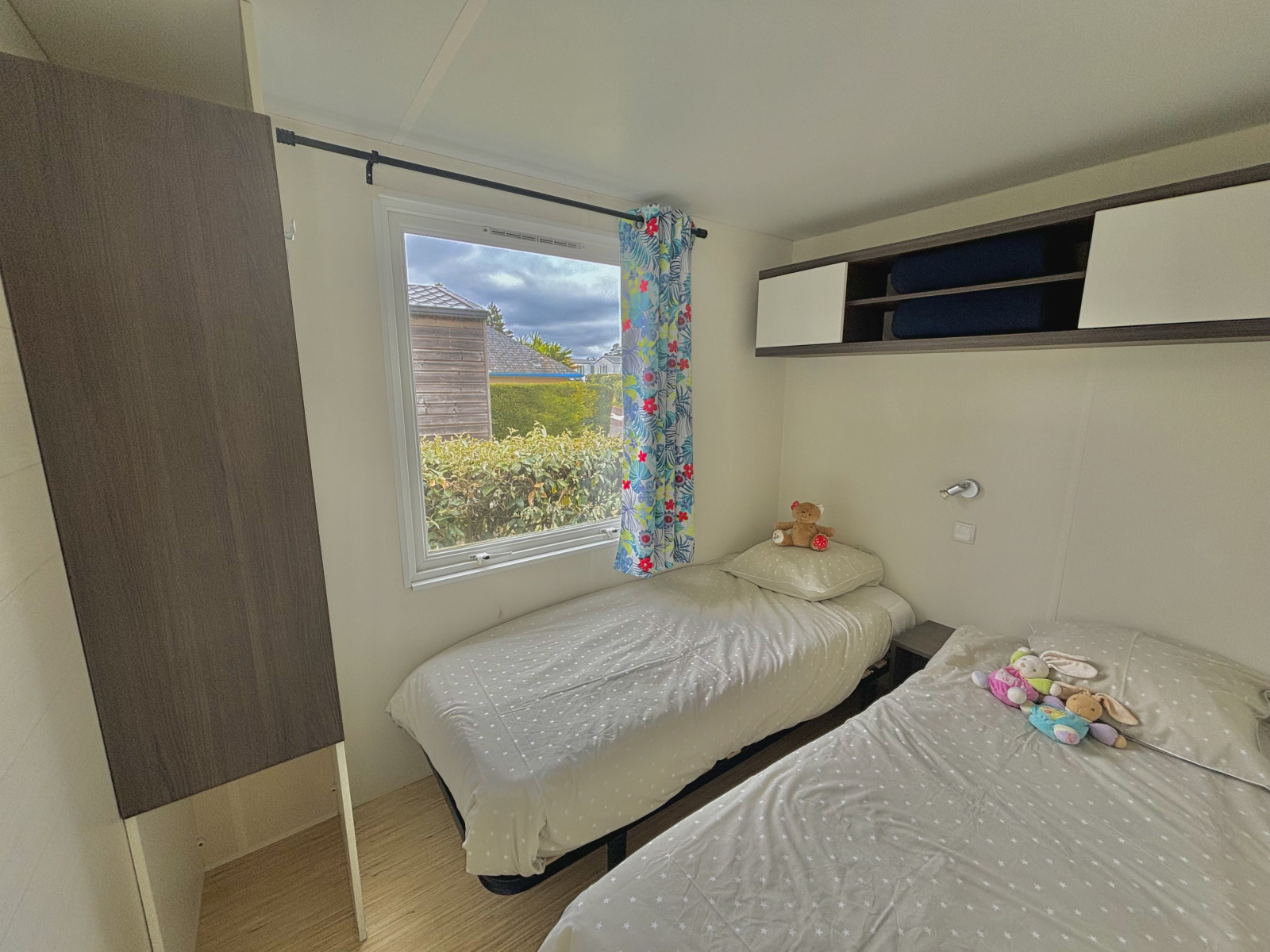 Knusse glampingkamer met twee eenpersoonsbedden, een bloemen gordijn en knuffels op de bedden.