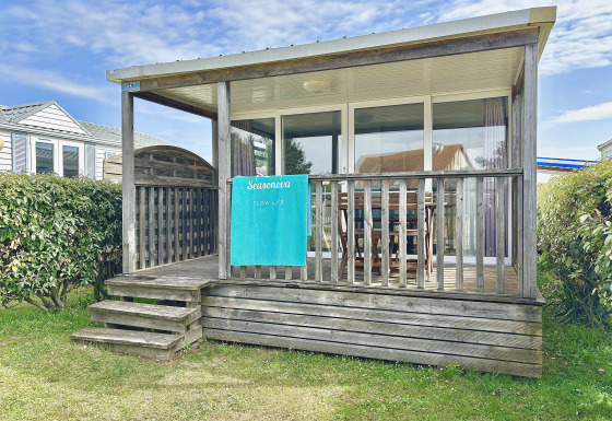 Cabaña de madera Cottage Zen con porche cubierto, puertas de cristal, toalla y setos verdes alrededor.