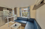 Vista interior de un alojamiento glamping con sofá azul en esquina, cocina pequeña y cortinas florales en la ventana.