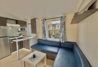 Vista interior de un alojamiento glamping con sofá azul en esquina, cocina pequeña y cortinas florales en la ventana.
