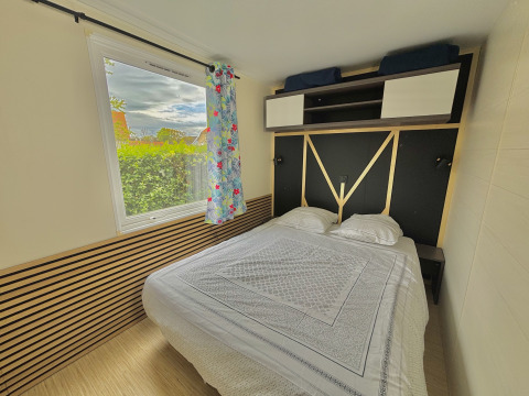 Chambre de glamping avec grand lit, placard en hauteur, vue sur la verdure et rideau fleuri.