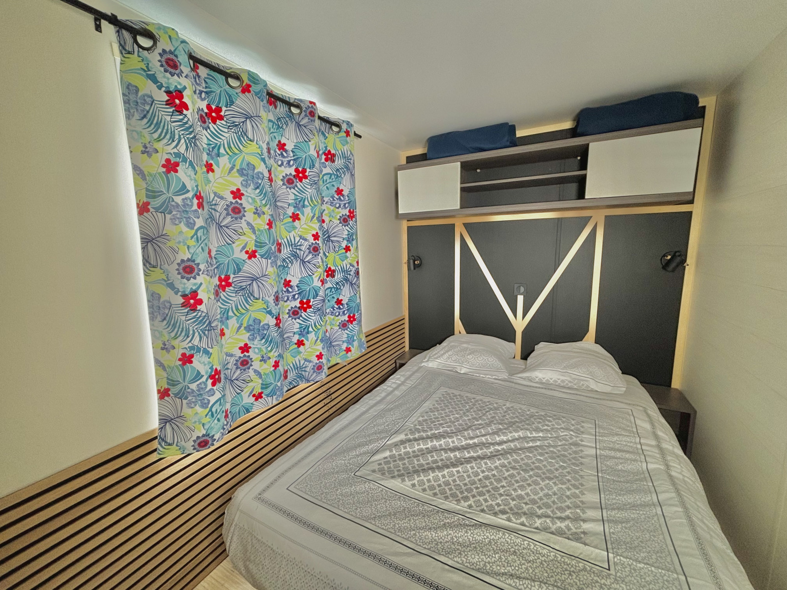 Slaapkamer in Cottage Zen bij Camping Seasonova Le Point du Jour, Frankrijk, met bloemen gordijnen en tweepersoonsbed.