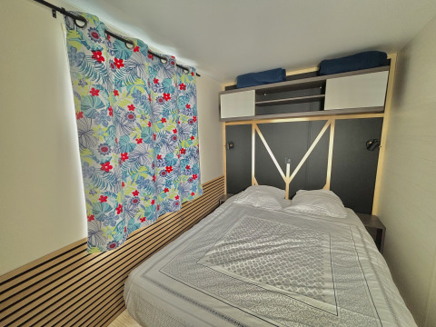 Schlafzimmer im Cottage Zen auf dem Campingplatz Seasonova Le Point du Jour, mit Blumenvorhang und Bett.