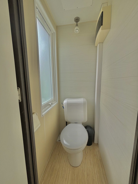 Baño compacto moderno con inodoro, ventana esmerilada y suelo símil madera en alojamiento glamping.
