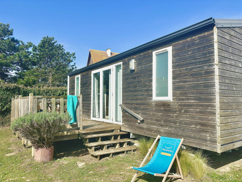 Cabaña de madera Cottage Evasion con terraza y tumbona bajo cielo azul, rodeada de vegetación.