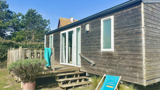Holzhaus Cottage Evasion mit Terrasse und Liegestuhl unter blauem Himmel, umgeben von grüner Natur.