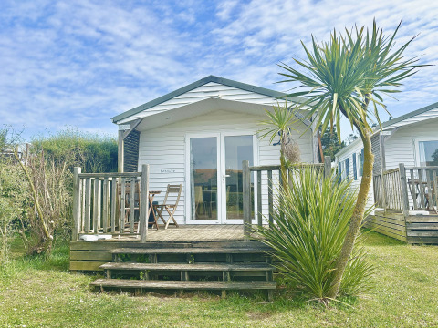 Cabaña blanca llamada Cottage Zen con terraza de madera, palmera y sillas bajo un cielo azul.