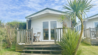 A bright white cabin named Cottage Zen with a porch, palm tree, and chairs, set under a blue sky.