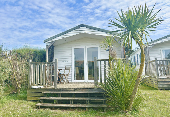 Cabaña blanca llamada Cottage Zen con terraza de madera, palmera y sillas bajo un cielo azul.