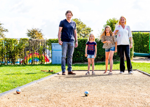 A family plays pétanque on a sunny court in a holiday park offering glamping accommodations.