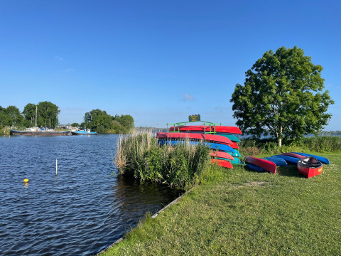 Kayaks et canoës au bord du lac dans un parc de vacances avec glamping, ciel bleu, arbres verts et bateaux.