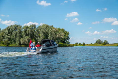 Menschen fahren mit einem kleinen Motorboot auf einem See bei einem Ferienpark mit Glamping-Angebot.