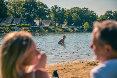 Famille au bord d’un lac dans un parc de vacances, hébergements glamping et chalets en arrière-plan.