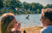 Familie nyder solen ved søbred med glampinghytter i baggrunden på en feriepark, sommerstemning.