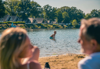 Family relaxes on sandy lakeshore at a holiday park, with glamping cabins and trees in the background.