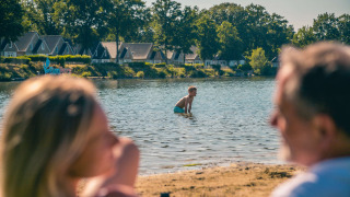 Familia descansa en la orilla de un lago en un parque vacacional con cabañas glamping y casas al fondo.