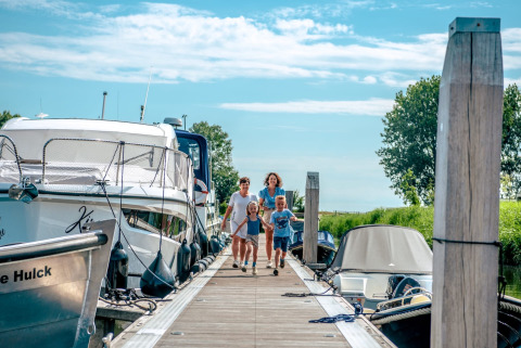 Gezin wandelt over een steiger tussen boten bij een vakantiepark met glamping, onder een blauwe lucht.