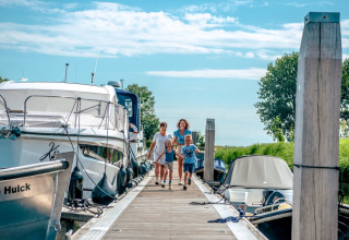 Famiglia che cammina su un pontile tra barche in un parco vacanze con glamping e cielo sereno.