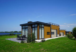 Moderna cabaña de glamping junto al agua en un parque vacacional, césped verde y zona de estar exterior.