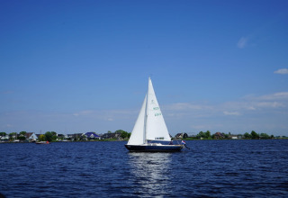 Zeilboot op het water bij een vakantiepark met glamping, huizen langs de oever en blauwe lucht op de achtergrond.