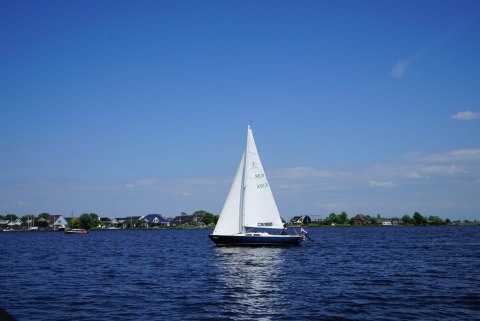 Velero navegando en un lago frente a un parque vacacional con glamping y casas cerca de la orilla al fondo.
