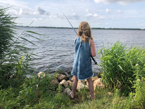 Mädchen im Jeanskleid angelt am Seeufer, umgeben von grünen Pflanzen, im Glamping-Ferienpark.