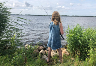 Pige i denimkjole fisker ved en sø omgivet af grønne planter i feriepark med glampingmuligheder.