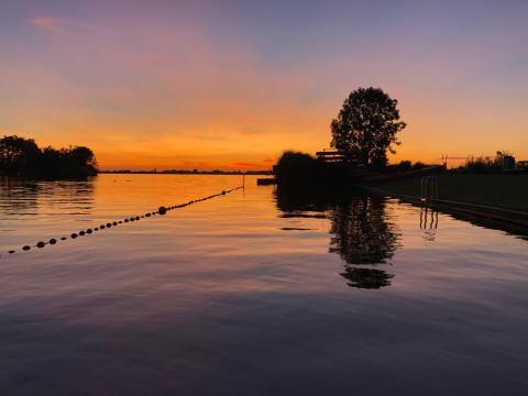 Zonsondergang over het water bij een vakantiepark met glamping, silhouetten van bomen en rustige sfeer.