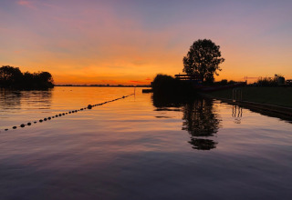 Zonsondergang over het water bij een vakantiepark met glamping, silhouetten van bomen en rustige sfeer.