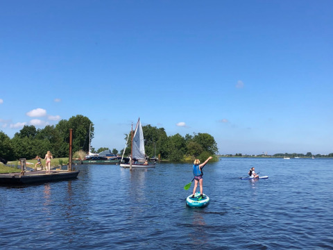 Holiday park with glamping; people paddleboard, sail, and enjoy the sunny lake and green scenery.