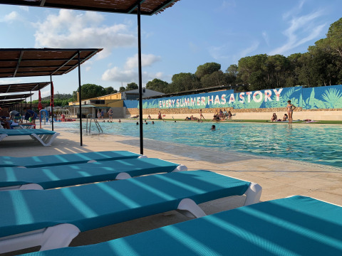 Piscina en Camping Mas Patoxas en Cataluña con tumbonas azules, toldos y gente disfrutando el verano.