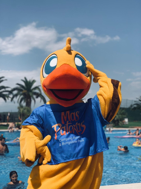Mascota con disfraz de pato del Camping Resort Mas Patoxas junto a la piscina con bañistas de fondo.