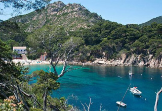 Scenic bay near Pals, Catalonia, with turquoise water, anchored boats, lush trees and mountains behind.