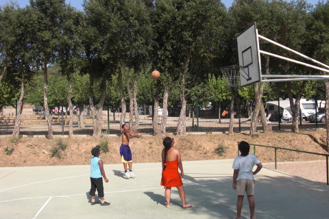 Niños juegan al baloncesto en una cancha al aire libre rodeada de árboles en Camping Mas Patoxas, Cataluña, España.