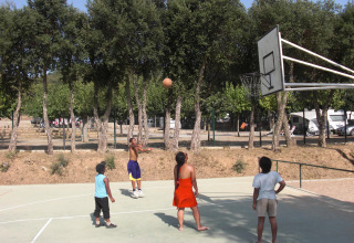 Kinderen spelen basketbal op een buitenplein omringd door bomen bij Camping Mas Patoxas in Catalonië, Spanje.