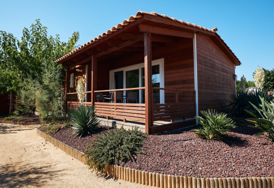 Chalet de madera Bungalow Quermany con terraza techada y jardinería de estilo desértico bajo un cielo despejado.