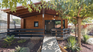 Chalet de madera Bungalow Quermany, con porche cubierto y rodeado de árboles y plantas de jardín.