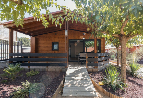 Chalet de madera Bungalow Quermany, con porche cubierto y rodeado de árboles y plantas de jardín.