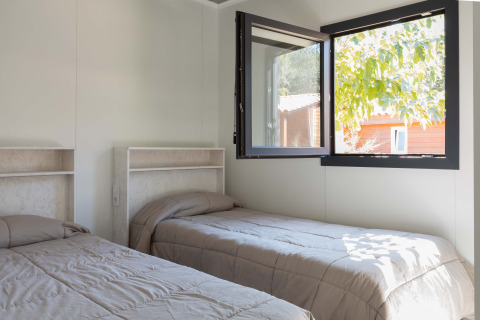 Dos camas individuales en una habitación luminosa con ventana abierta en Bungalow Quermany, Camping Mas Patoxas, España.