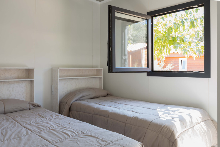 Twee eenpersoonsbedden in een lichte kamer met open raam in Bungalow Quermany, Camping Mas Patoxas, Spanje.