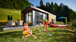Two people enjoy a barbecue on the grass outside a modern Panorama Chalet surrounded by lush nature.