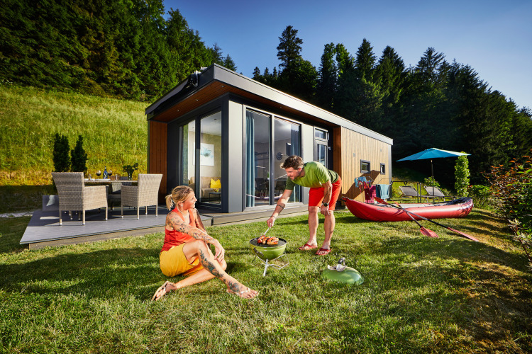 Dos personas disfrutan de una barbacoa en el césped junto a un moderno Panorama Chalet rodeado de naturaleza.