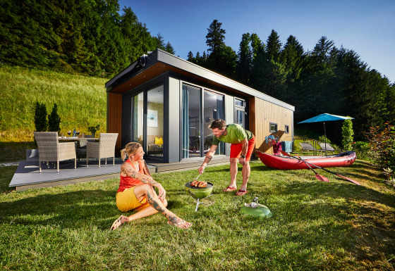 Two people enjoy a barbecue on the grass outside a modern Panorama Chalet surrounded by lush nature.