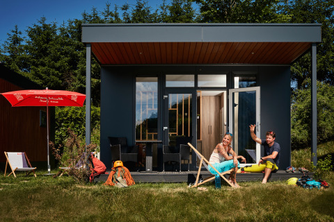 Twee personen relaxen voor een tiny house genaamd Studio, met tuinstoelen en avonturenmateriaal.