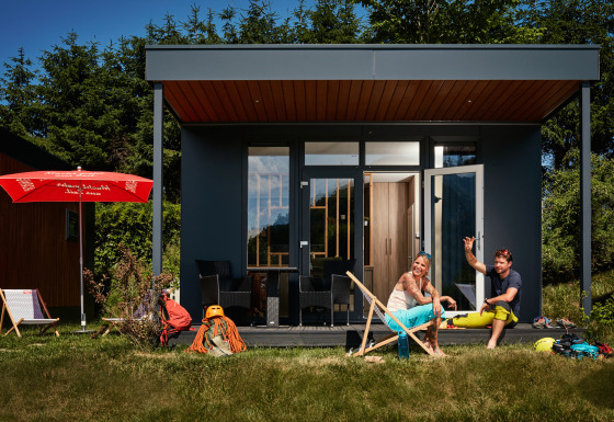 Dos personas se relajan frente a una tiny house llamada Studio, con sillas y equipo de aventura en el césped.