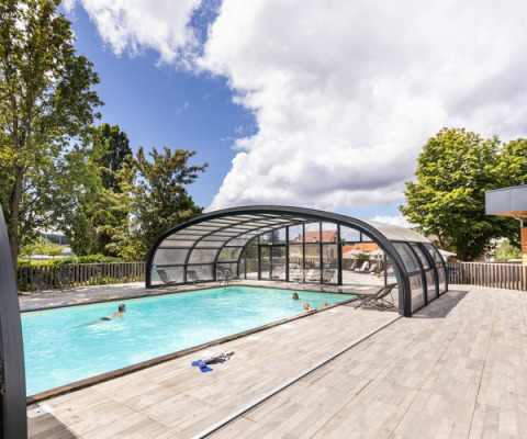 Piscina exterior con cubierta de vidrio y visitantes relajándose en Huttopia Versailles, Île de France, Francia.