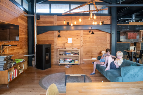 Moderne træhytte loungeområde i Huttopia Versailles med børn på sofa, bøger og industrielle detaljer.