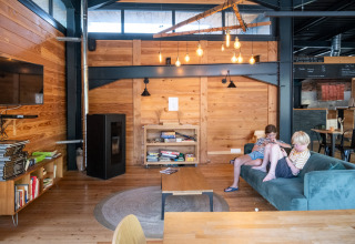 Moderne træhytte loungeområde i Huttopia Versailles med børn på sofa, bøger og industrielle detaljer.